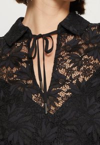 Chemise en dentelle noire avec des motifs floraux, dotée d'un col et d'un détail à nouer au niveau de l'encolure, mettant en valeur une texture complexe et un design en couches.
