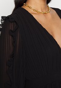 Femme portant une robe noire transparente à manches volantées avec des plis et un collier de perles dorées autour du cou.
