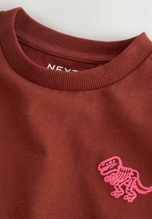 Brown shirt with a round neckline featuring a pink dinosaur skeleton graphic on the front. The fabric appears soft and casual.