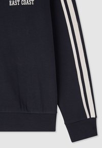 Manica di felpa blu navy con due strisce bianche e polsino a costine, parte del testo "EAST COAST" visibile sul tessuto superiore.