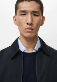 Chaqueta negra con cremallera y cuello puntiagudo sobre un suéter azul marino y una camisa de rayas azul claro. Tejido suave y diseño ajustado.