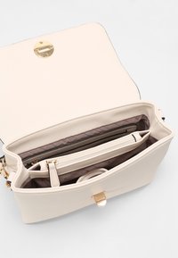 Borsa a tracolla in pelle beige con una chiusura circolare dorata. L'interno presenta una fodera trapuntata, una tasca con zip e diversi scomparti per l'organizzazione.