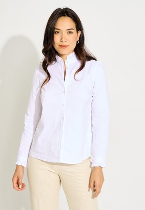 Mujer con el pelo largo y oscuro, que lleva una camisa blanca abotonada con cuello y puños con volantes, y pantalones beige acanalados, de pie frente a un fondo liso.