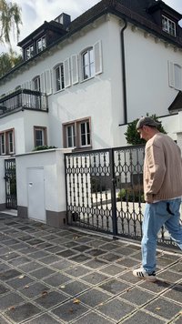 Muž v béžovom svetri, modrých džínsoch a šiltovke kráča okolo čiernej kované železnej brány pred bielym viacposchodovým domom so žalúziovými oknami.