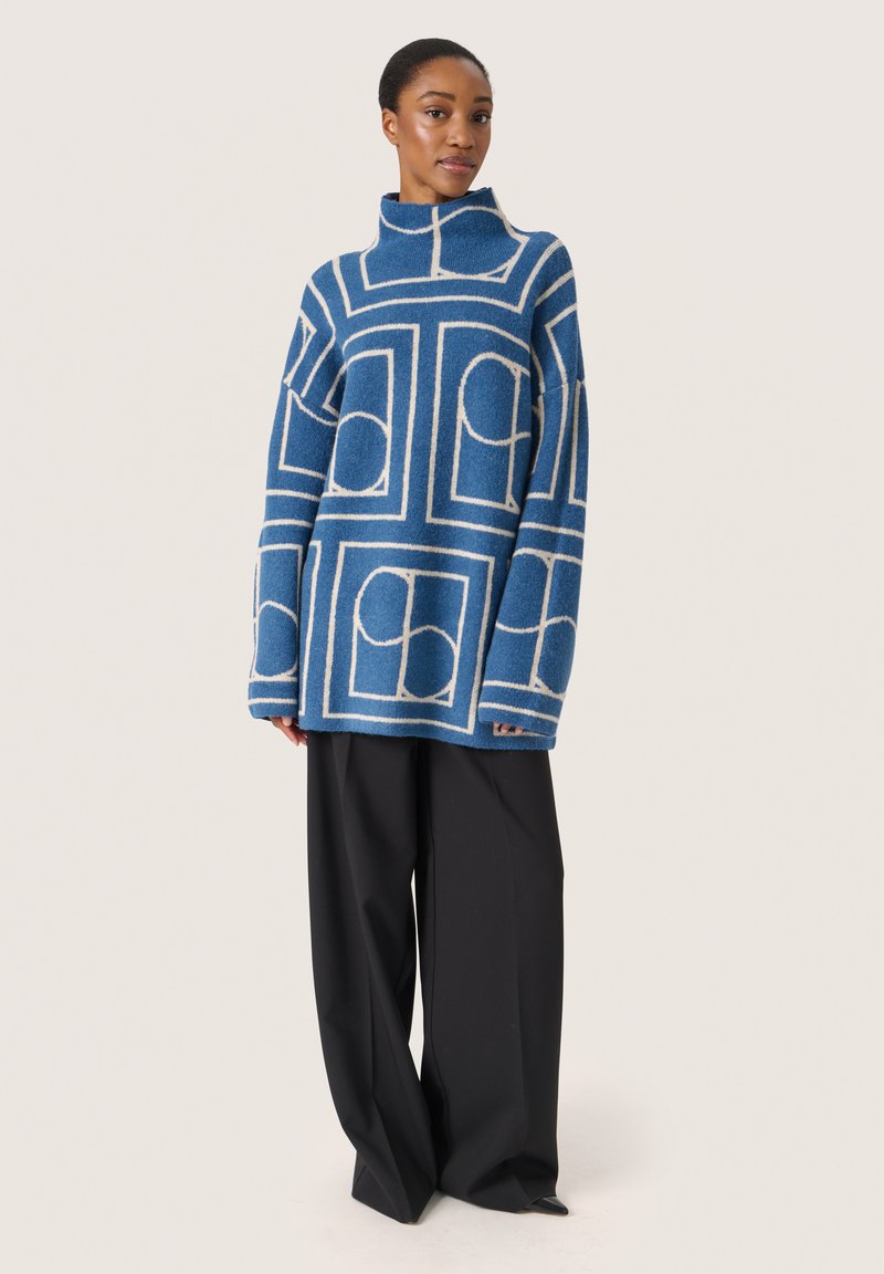 Blauer oversized Pullover mit geometrischem weißen Muster, hoher Kragen und lockerer Passform, kombiniert mit weiten schwarzen Hosen.