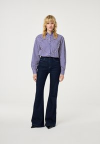 Vrouw met blond haar, gekleed in een paars-witte gestreepte blouse met een gerimpeld hartontwerp en donkere flared jeans, staande voor een witte achtergrond.