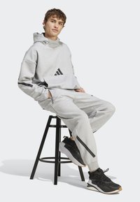 Sivá mikina s čiernymi akcentmi a zipsami; v kombinácii so zodpovedajúcimi sivými teplákmi a čiernymi športovými topánkami. Model sedí na čiernej stoličke.