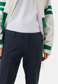 Pantaloni navy con chiusura a bottone e passanti per cintura, indossati con una t-shirt bianca e una giacca con zip dai dettagli verdi e strisce.