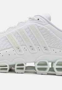 Scarpa sportiva bianca con tomaia in rete, tre accenti a strisce distintive e un suola imbottita unica con ritagli arrotondati.