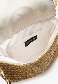 Vevd stråveske med gullkjede, beige innerside og inn pockets med en svart merkeetikett.