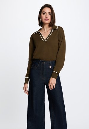 Frau mit kurzen braunen Haaren, die einen olivgrünen Langarmpullover mit weißem Rand und dunkle, weit geschnittene Jeans trägt, steht vor einem weißen Hintergrund.