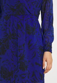 Blå blommig klänning med ett svart blommönster, långa ärmar, elastiska manschetter och en åtsittande midja med veckdetalj.