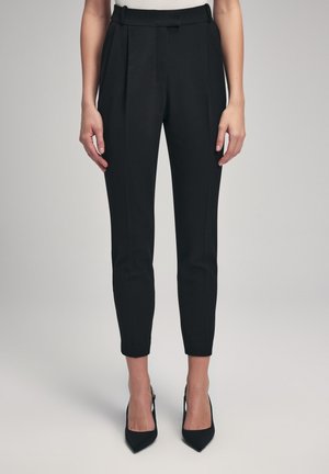 Black tailored trousers with a high waistband, featuring pleats, a slim fit, and cropped length. Worn with pointed black heels.