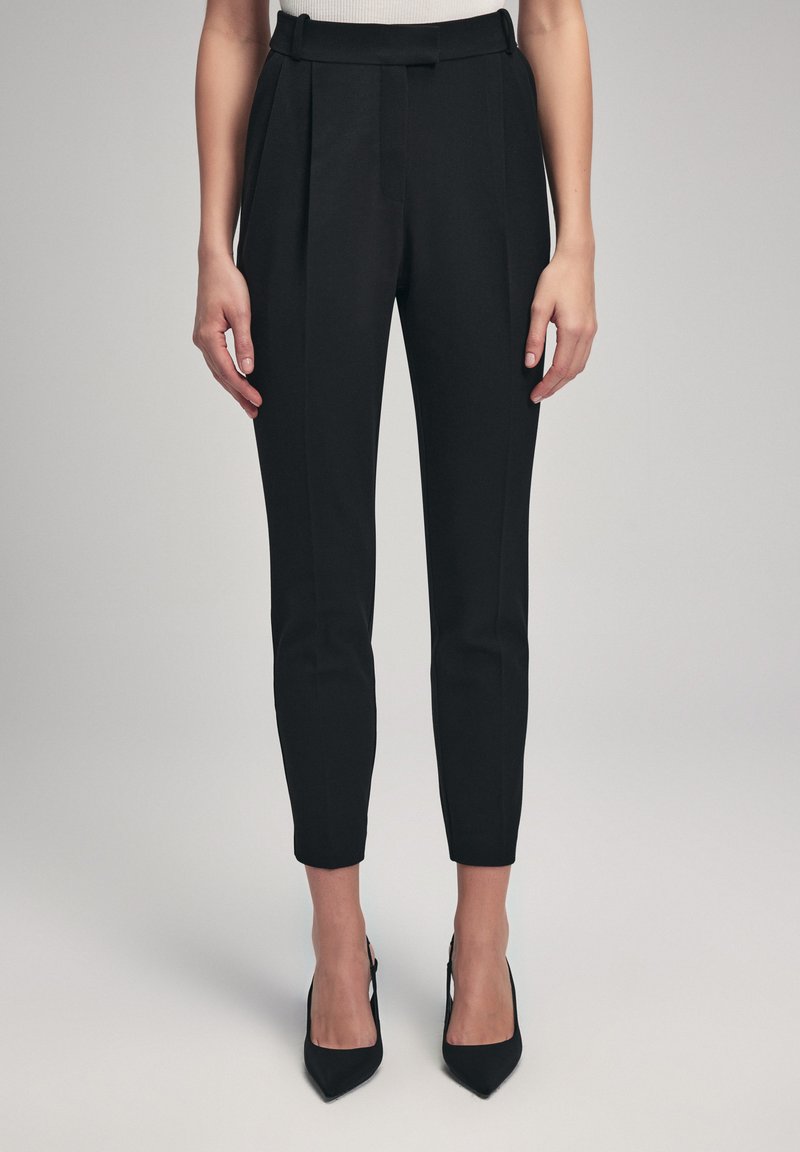 Zwarte op maat gemaakte broeken met een hoge tailleband, met plooien, een slim fit en een verkorte lengte. Draagt zwarte spitse hakken.