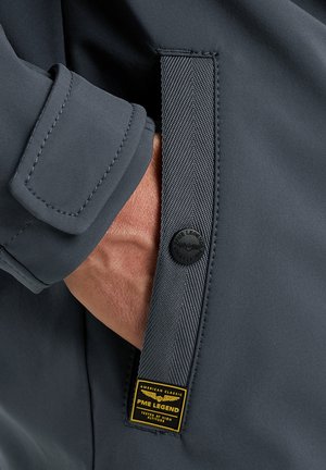 Roka delno v žepu temno sive jakne z zašito zanko, črnim sponkom in rumeno nalepko PME Legend.