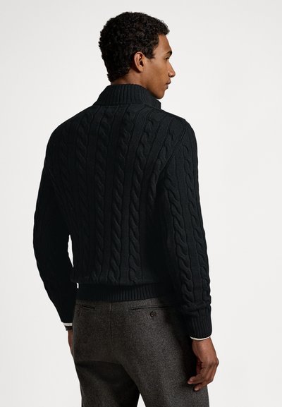 Μαύρο πλεκτό πουλόβερ με ψηλό γιακά, διαθέτει σχέδιο με πλεξούδα, ριμπ μανσέτες και περιλαίμιο. Φορεμένο πάνω από γκρι παντελόνια.