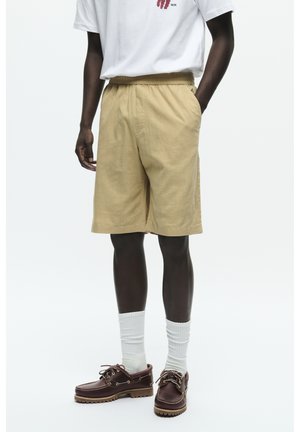 Shorts caqui con cinturilla elástica, hasta la rodilla, con bolsillos laterales. Combinados con calcetines blancos y zapatos náuticos marrones.