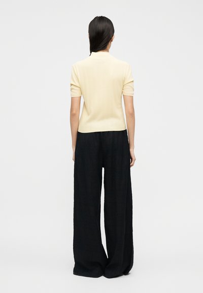 Γυναίκα με μακριά σκούρα μαλλιά που στέκεται με την πλάτη, φοράει ανοιχτόχρωμο τοπ με κοντό μανίκι και φαρδιά μαύρη παντελόνα.