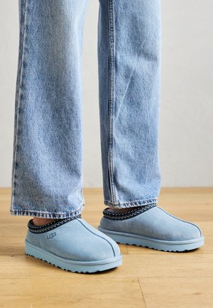 Chaussons bleu clair à enfiler avec bordure noire à motifs, portés avec un jean bleu droit sur un sol en bois.