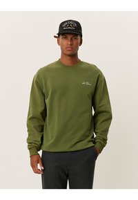 Felpa verde in cotone con maniche lunghe, collo a giro e un piccolo logo ricamato sul petto. Indossata con pantaloni scuri e un berretto.
