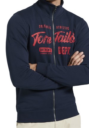 Mężczyzna z założonymi skrzyżowanymi ramionami, ubrany w granatową bluzę rozpinaną na zamek z czerwonym napisem „Tom Tailor AUTHENTIC DEPT.” na piersi, widoczne beżowe spodnie poniżej.