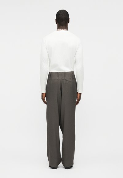 Άνδρας που φοράει άσπρο πουκάμισο με μακριά μανίκια και ψηλόμεσα, φαρδιά σκούρα γκρι παντελόνια, στέκεται γυρισμένος προς τα πίσω σε απλό φόντο.