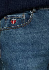 Denim jeans hebben een middelblauwe kleur met subtiele vervaging. Een patch met "Scholk" in marineblauw en rood is op de voorkant van de zak genaaid.
