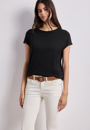 Kobieta ubrana w czarną bluzkę z krótkim rękawem, jasnobeżowe spodnie i brązowy pasek, stojąca na tle jednolitego jasnoszarego tła.