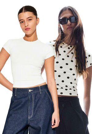 To modeller viser bluser: en figursyet hvid t-shirt med korte ærmer og en sort t-shirt med hvide prikker og korte ærmer, kombineret med mørk denim og sorte bukser.