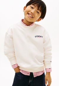 Λευκό φούτερ με ριμπ μανσέτες, με την επιγραφή "TOMMY HILFIGER" σε ναυτικό μπλε. Φορεμένο πάνω από ένα κόκκινο και άσπρο ριγέ πουκάμισο με γιακά, και μπλε τζιν παντελόνι.