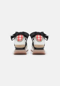 Sportovní boty s bílou koženou svrškem, černými akcenty, červeným kostkovaným detailem na patě a tkanými prvky v béžové barvě. Na zadní straně mají velké černé mašle.