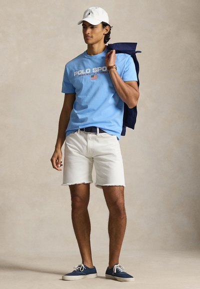 Ανοιχτό μπλε βαμβακερό μπλουζάκι με γραφικό "POLO SPORT", λευκά τζιν σορτς, μπλε ναυτικό αθλητικά παπούτσια και λευκό καπέλο μπέιζμπολ, κρατώντας ένα μπλε ναυτικό σακάκι.