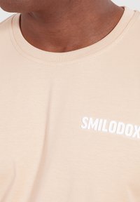 Lichtbeige t-shirt met een ronde hals en korte mouwen, met "SMILODOX" in het wit gedrukt op het linker borstgebied.