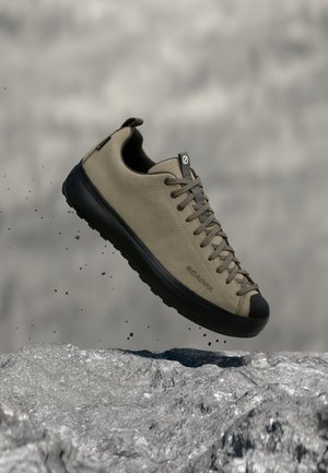 Hellbrauner Sneaker mit schwarzer Sohle und Schnürsenkeln, der über felsigem Boden schwebt, wobei kleine Steine um ihn herumfallen.