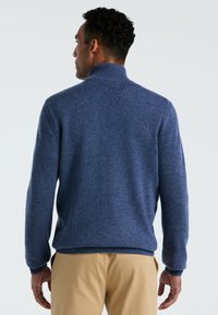 Blauwe gebreide trui met een hoge kraag, geribde manchetten en zoom. Zachte textuur en een effen kleur, van achteren gezien, gecombineerd met beige broek.