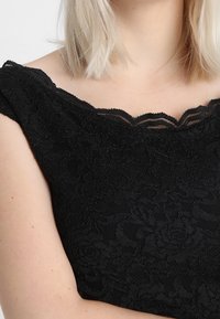 Svart spetsklänning med en scallopad halsringning och blommönster, med korta ärmar och en figurnära bodice som framhäver den texturerade tyget.