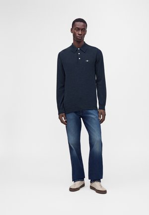 Homme portant un polo à manches longues foncé, un jean bleu et des baskets beiges, debout devant un fond clair uni.