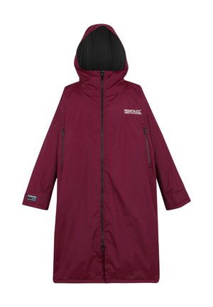 Mężczyzna w długiej bordowej kurtce Regatta z pomarańczowym polarem w środku, czarnych spodniach, białych trampkach i czarnej czapce, pozujący na jednolitym tle.