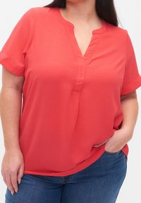Blouse corail avec un col en V, manches courtes et tissu texturé. Associée à un jean bleu, à coupe décontractée et avec des fentes latérales.