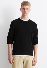 Schwarzer gestrickter Pullover mit einem strukturierten Design, Rundhalsausschnitt und gerippten Bündchen, über einem weißen Hemd getragen. Kombiniert mit hellbeigen Hosen.