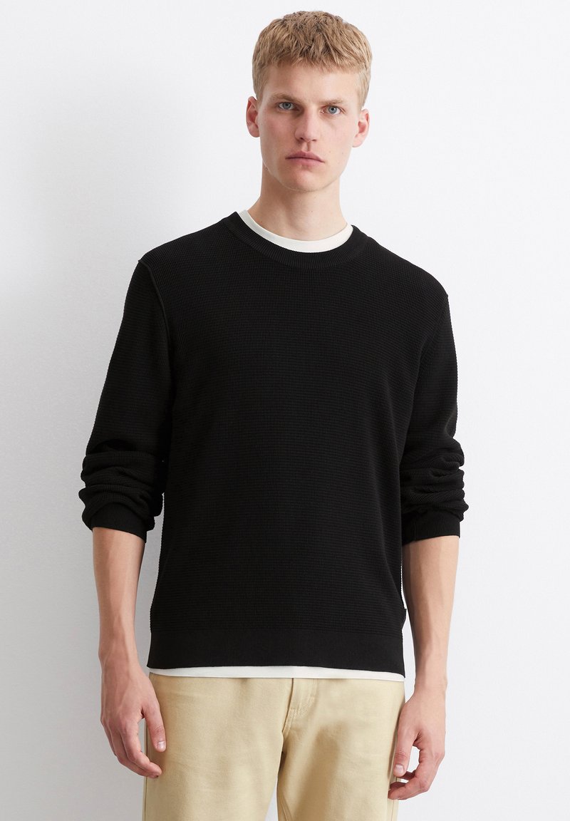 Schwarzer gestrickter Pullover mit einem strukturierten Design, Rundhalsausschnitt und gerippten Bündchen, über einem weißen Hemd getragen. Kombiniert mit hellbeigen Hosen.