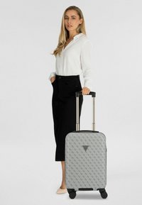 Mujer con blusa blanca y falda negra de pie junto a una maleta gris con patrón y asa extendida sobre fondo blanco.