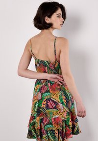 Eine junge Frau mit kurzen Haaren trägt ein farbenfrohes, gemustertes Kleid mit einem Rüschensaum und steht vor einem schlichten Hintergrund.