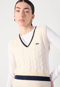 Cremfarbener, kabelgestrickter Pullunder mit V-Ausschnitt, der marineblaue und gelbe Streifen am Saum aufweist und ein kleines grünes Logo auf der linken Seite hat.