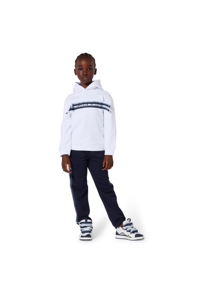 Bambino in piedi che indossa una felpa con cappuccio bianca con striscia blu, pantaloni blu scuro con strisce laterali e scarpe da ginnastica bianche con lacci spessi e decorati.