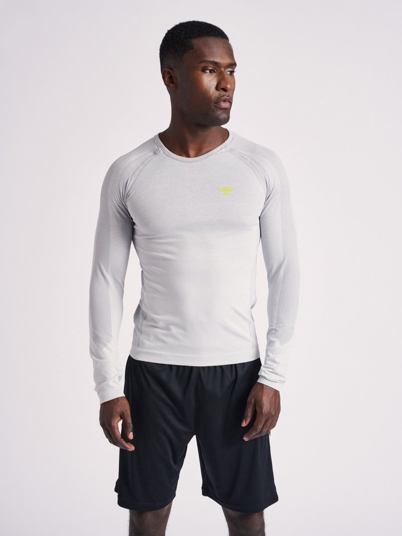 Homme debout, portant un t-shirt de sport gris cintré à manches longues avec un petit logo jaune et un short noir, regardant vers sa droite.