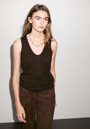 Top nero senza maniche in maglia con scollo a V, abbinato a pantaloni scamosciati marroni. La modella indossa una collana in argento. Design semplice e pulito.