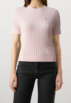 Kvinde iført en lyserød kortærmet textured striktop og sorte højtaljede jeans, stående med armene afslappet ned langs siden.