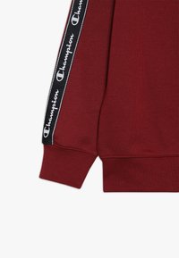 felpa borgogna con orlo e polsini a coste, dotata di strisce laterali nere con il logo "Champion" in lettere bianche.