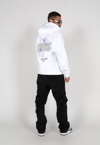 Weißer Hoodie mit einem Schmetterlingsgrafik und Text auf dem Rücken, kombiniert mit schwarzen Cargo-Hosen und Sonnenbrille, vor einem einfachen Hintergrund.